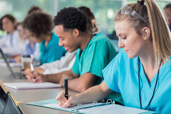 Diverse male and female young medical students confidently take notes during their professor's lecture.They are wearing scrubs.